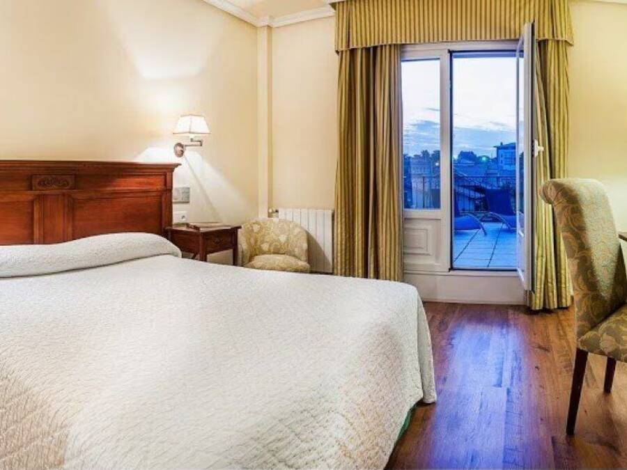 A hotel room with a large bed, a desk, a chair, and a view of the city through a window.
