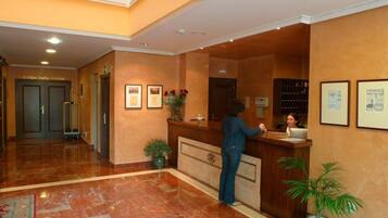 A hotel reception area with a receptionist, a reception desk, framed pictures on the wall, and a potted plant.