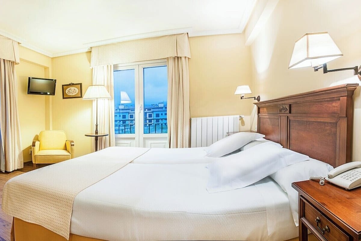 A hotel room with a large bed, a chair, a desk with a phone, a TV, and a window with curtains.