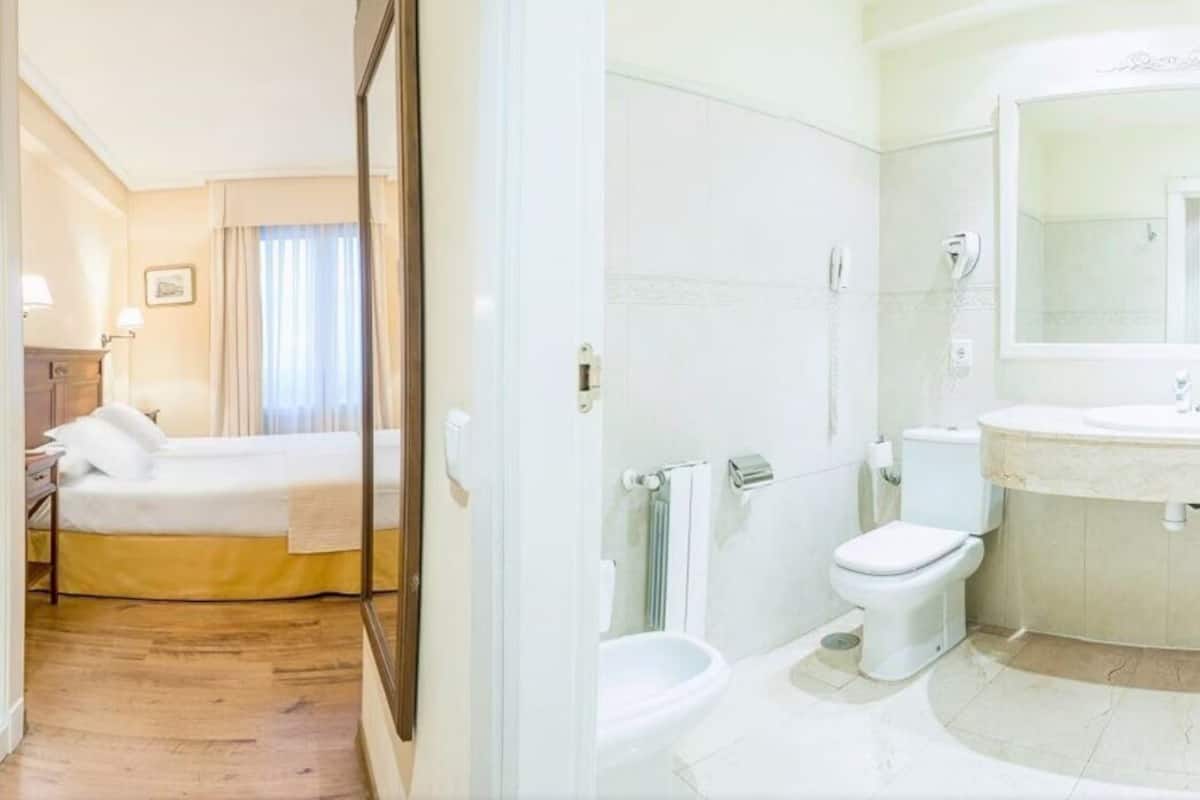A hotel room with a bed, a bedside table, a mirror, and a bathroom with a toilet, bidet, and sink.