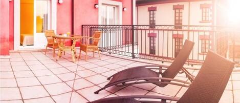 Un balcón con sillas de mimbre y una mesa, un edificio rojo y un edificio blanco con balcón.
