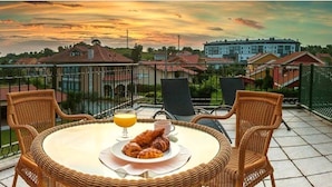 Un balcon avec des fauteuils en osier, une table ronde, des croissants, un verre de jus d’orange et une vue sur les bâtiments en arrière-plan.