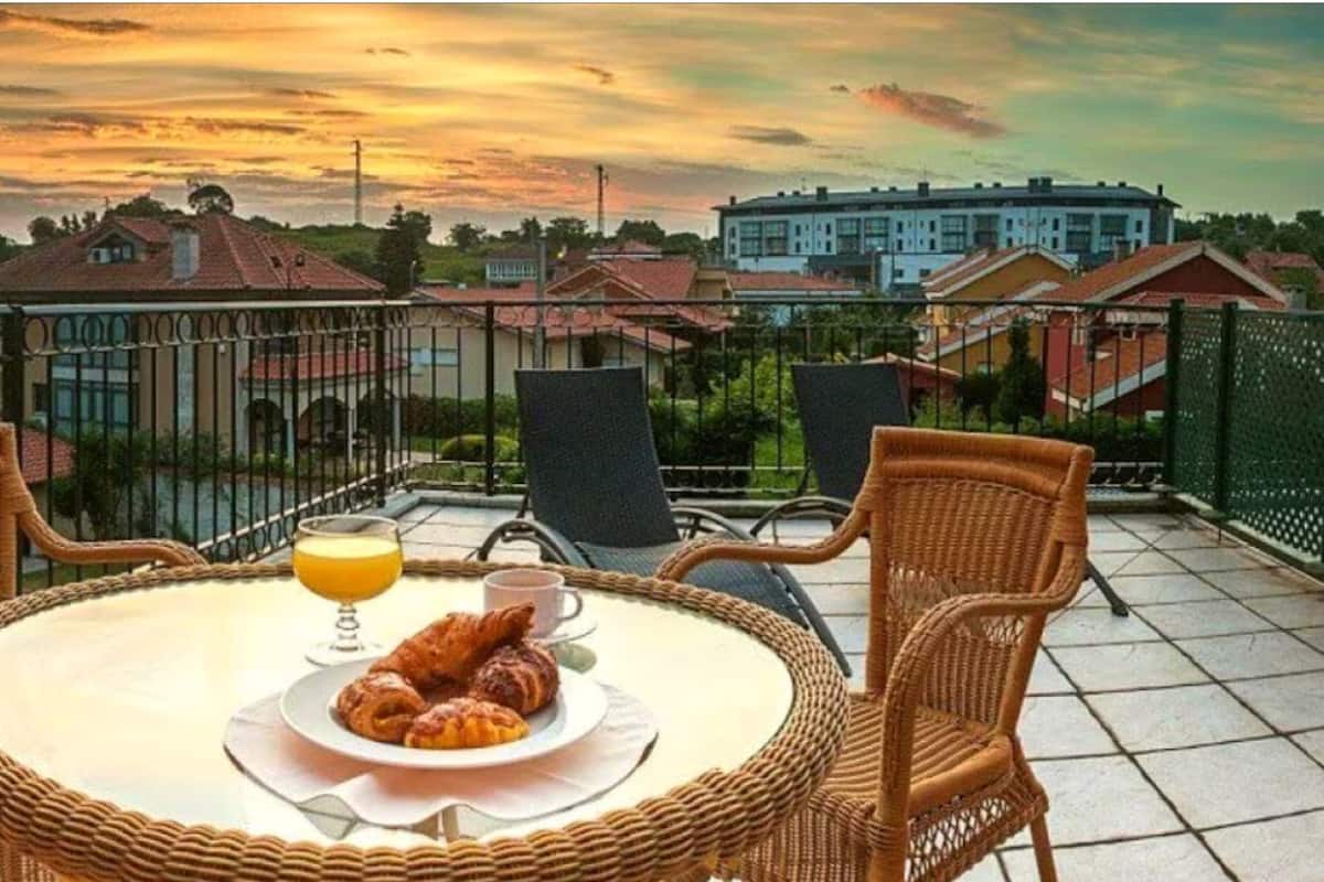 A balcony with wicker chairs, a round table, croissants, a glass of orange juice, and a view of buildings in the background.