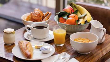 Una mesa de desayuno con cruasanes, un cuenco de fruta, un vaso de jugo de naranja, una taza de café, una jarra de leche y un tazón de cereales.