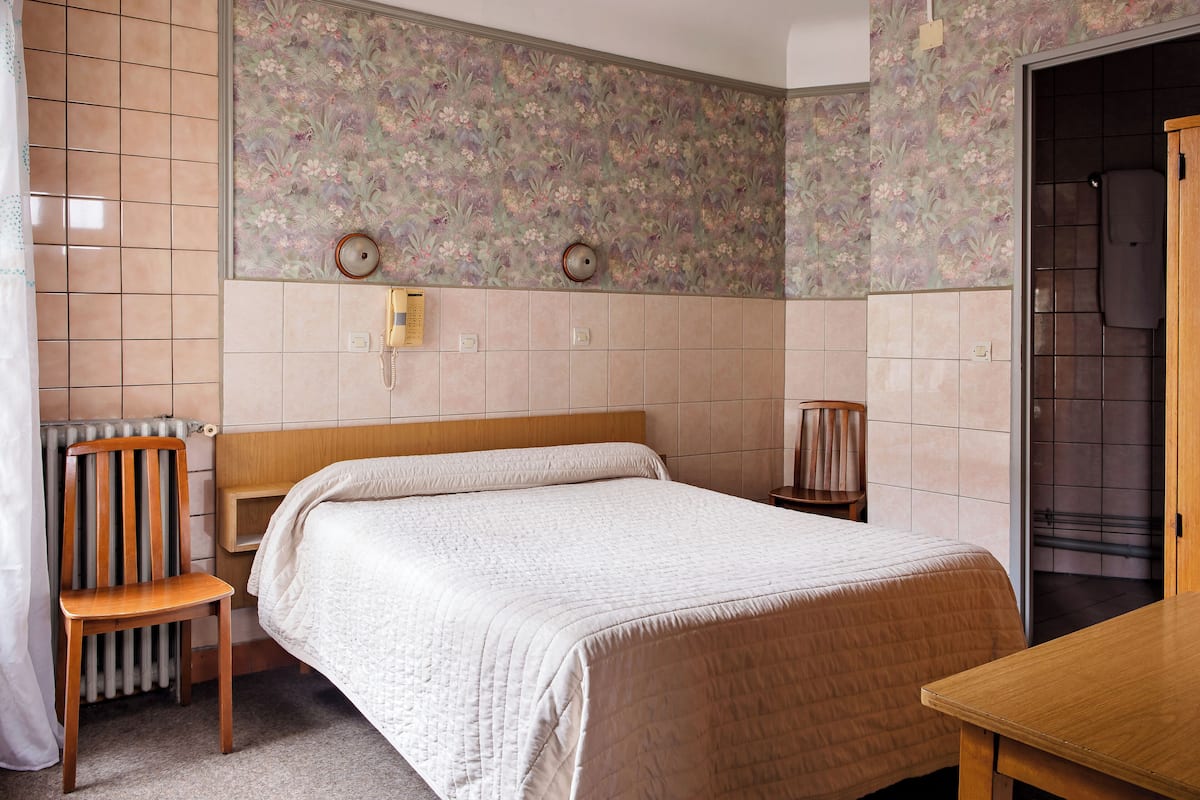 A bedroom with a wooden headboard, a single bed with a quilt, a chair, a radiator, and a bathroom visible through an open door.