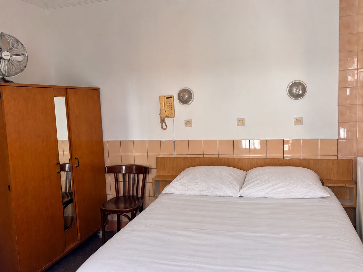 Habitación de hotel con cama, armario de madera, silla, teléfono y lámpara de pared.