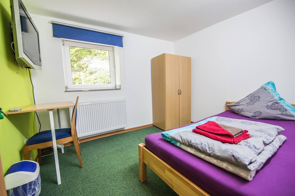 Ein kleiner, ordentlicher Raum mit einem Bett, einem Holzschrank, einem Schreibtisch und einer Heizung.