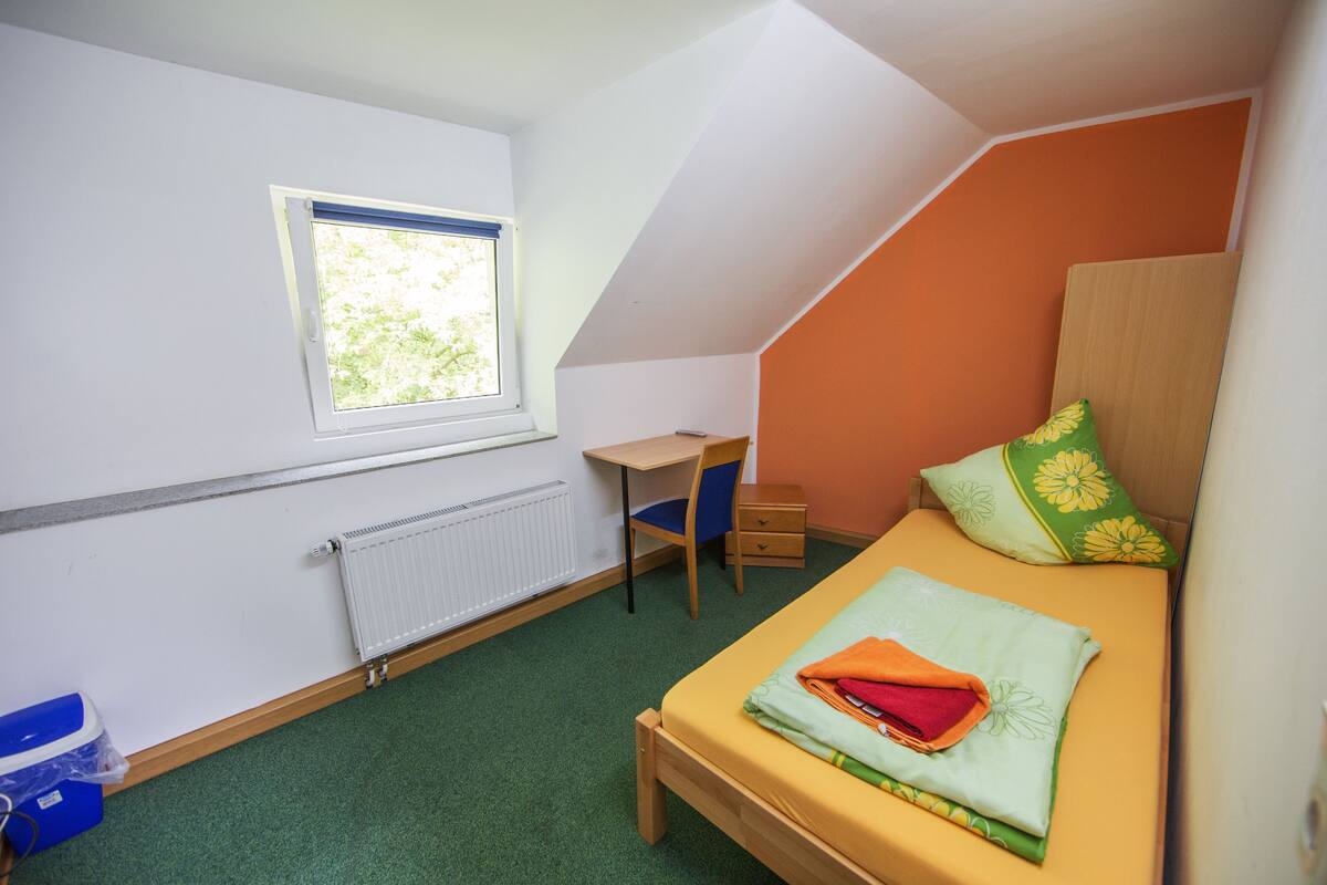 Ein kleiner, gut beleuchteter Raum mit einem Einzelbett, einem Schreibtisch und einem Fenster.