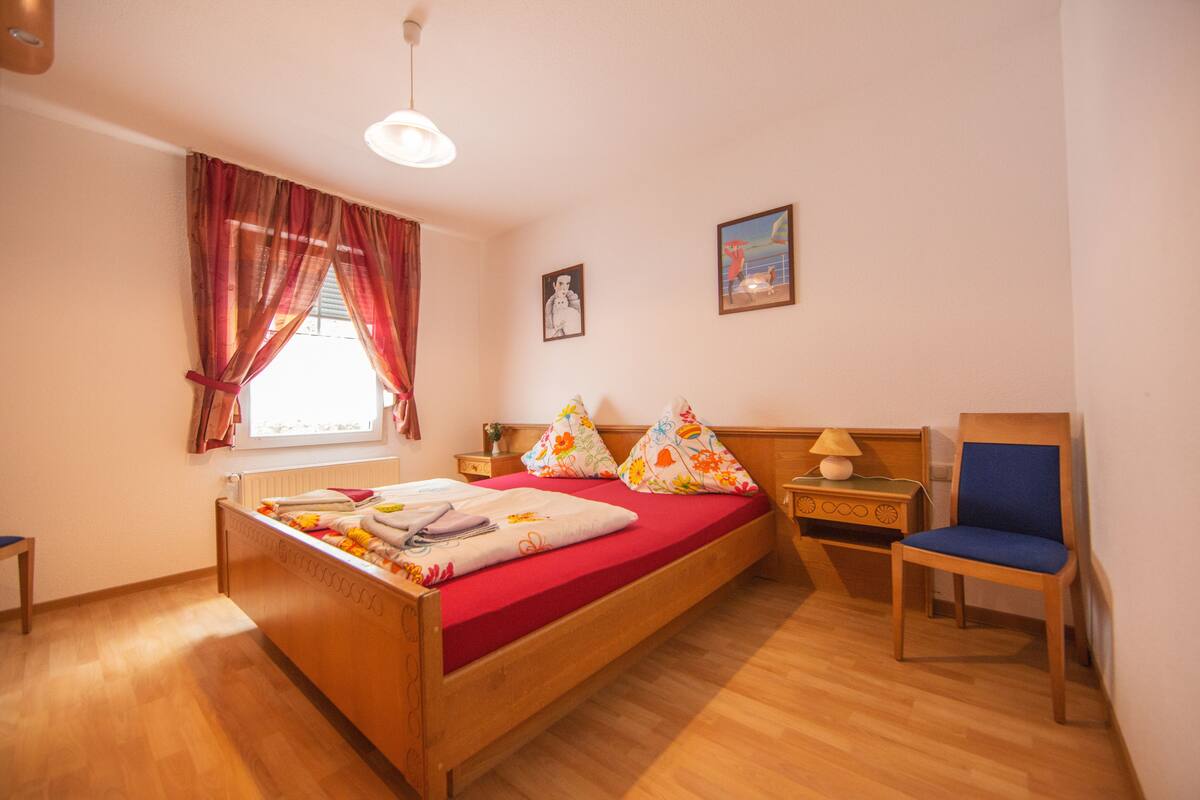 Ein Schlafzimmer mit einem hölzernen Bett, roten Vorhängen, einem Holzstuhl und einem Nachttisch mit Lampe.
