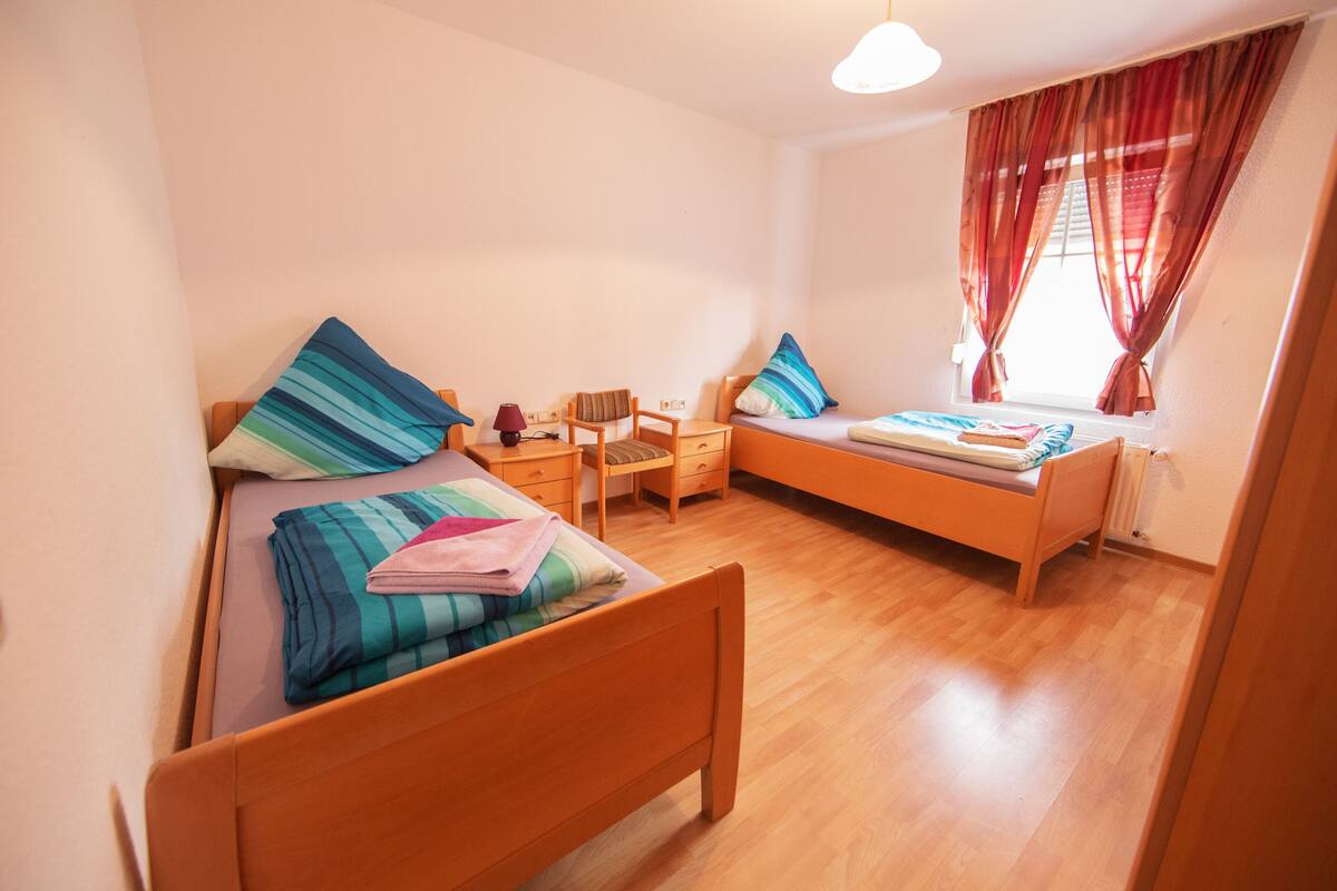 Ein kleines, gut beleuchtetes Zimmer mit zwei Einzelbetten, einem Holztisch, einem Stuhl und einem Fenster mit Vorhängen.