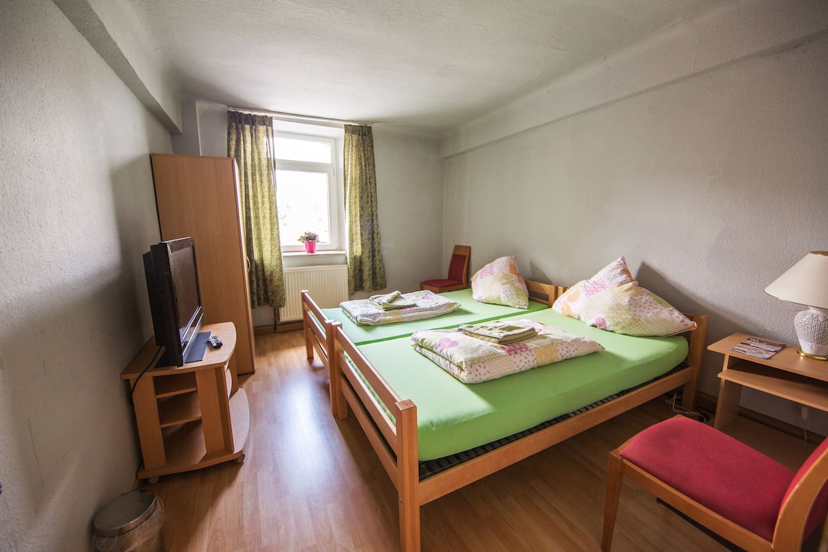 Ein kleines, ordentliches Zimmer mit einem Einzelbett, einem Holzschrank, einem Fernseher auf einem Standfuß, einem kleinen Tisch mit einer Lampe und einem Fenster mit Vorhängen.