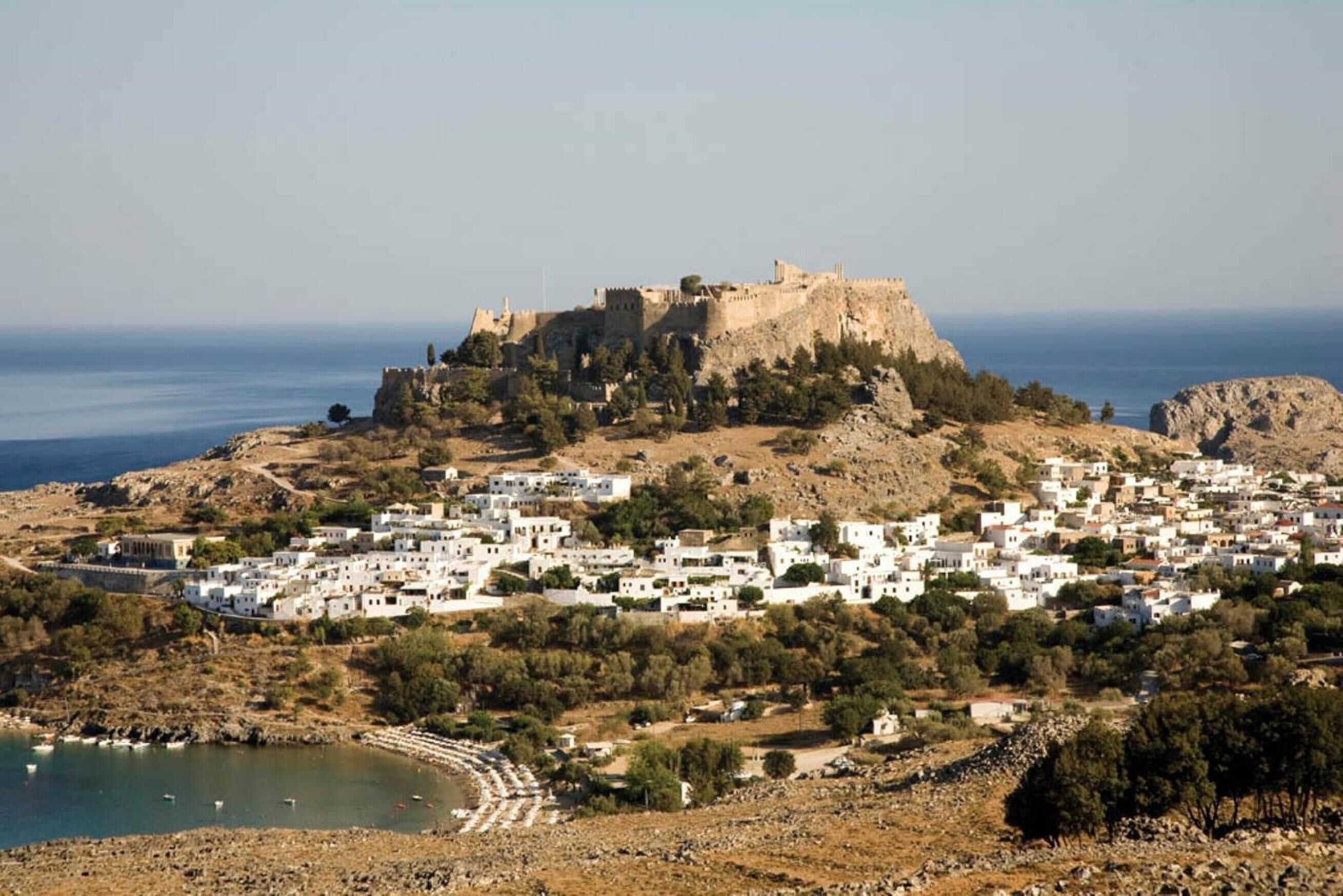 Un pueblo costero con un castillo destacado en una colina, rodeado de edificios blancos y una pequeña playa.