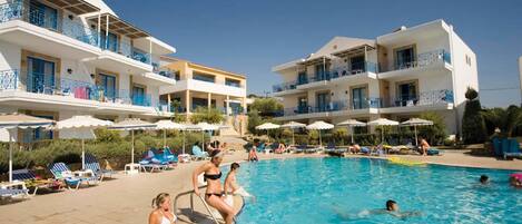 A swimming pool area with multiple buildings, sun loungers, and umbrellas.