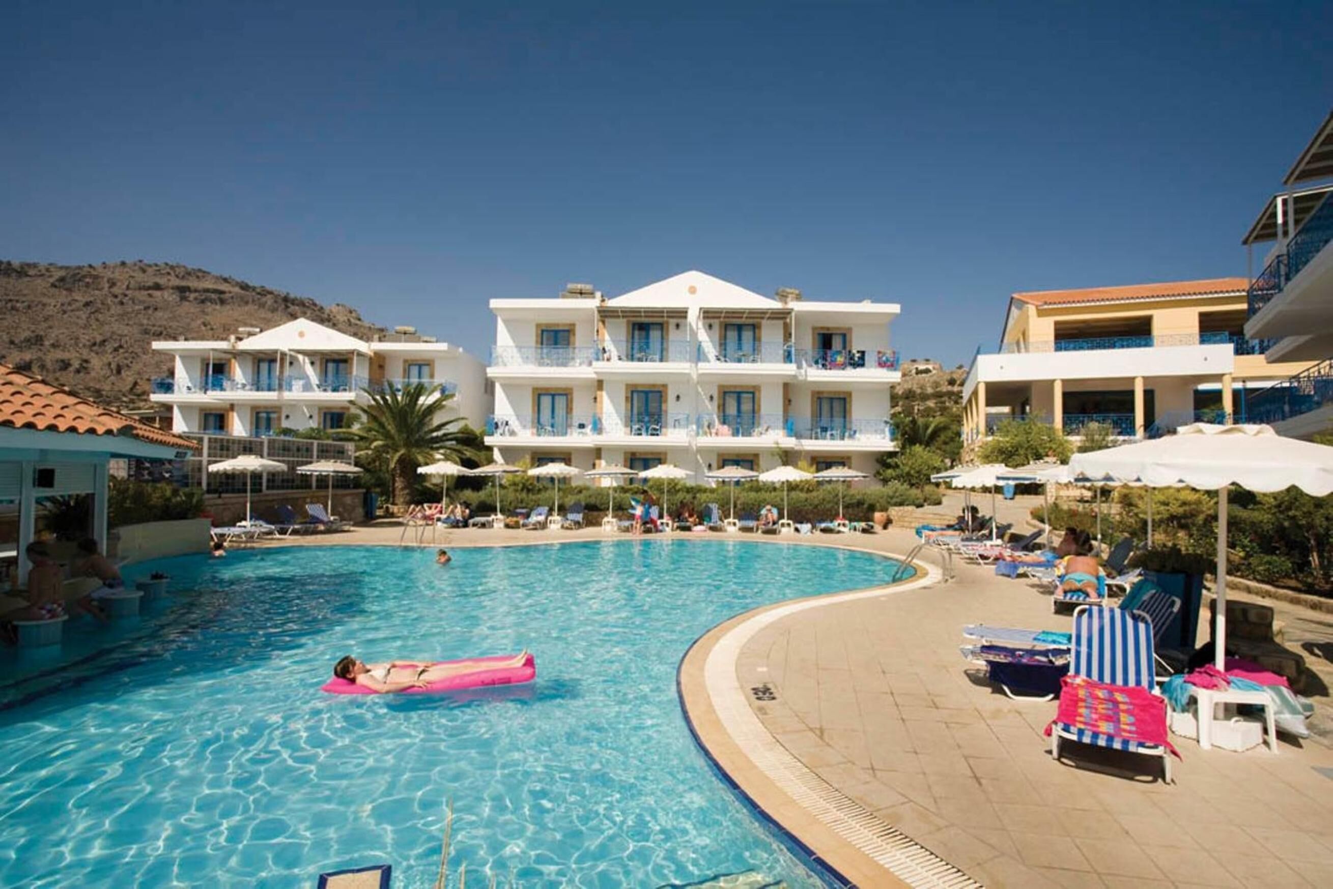 Une piscine avec des chaises longues et des parasols, un bâtiment doté de balcons, et un paysage montagneux en arrière-plan.