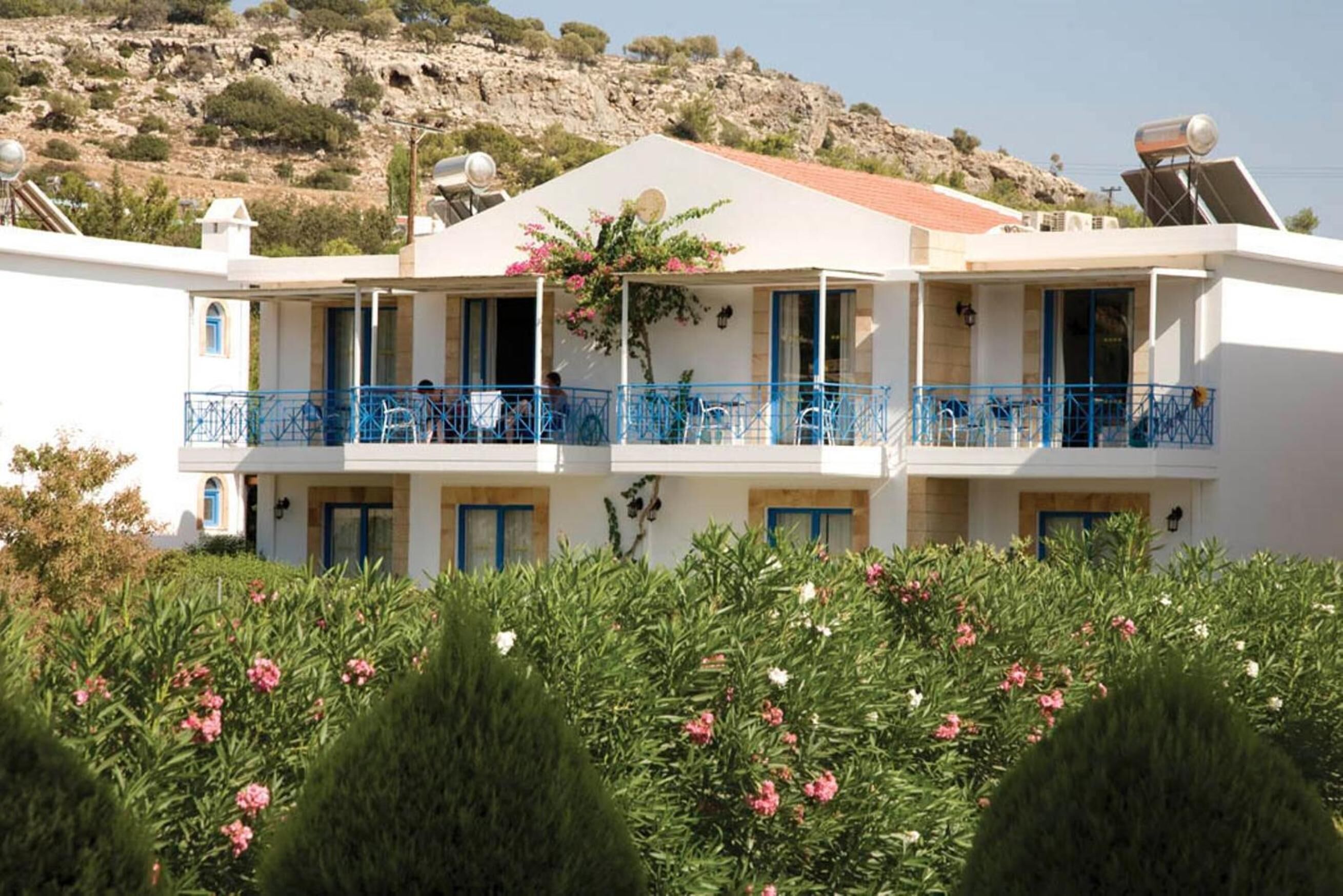 Un edificio de dos pisos con balcones, un paisaje montañoso de fondo y un jardín con flores rosadas.