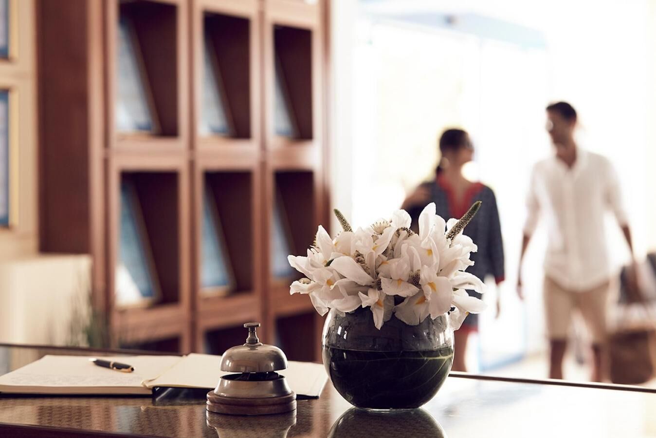 A hotel reception desk with a vase of white flowers, a pen, and a bell on a polished surface.
