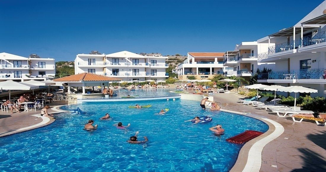 A swimming pool surrounded by white buildings with balconies, sun loungers, and umbrellas.