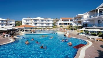 A swimming pool surrounded by white buildings with balconies, sun loungers, and umbrellas.