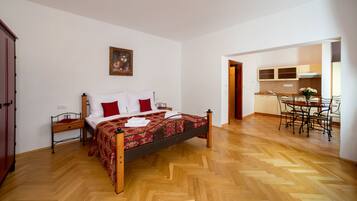 Een slaapkamer met een houten bed, een nachtkastje, een rood kussen en een schilderij aan de muur. Er ligt een houten vloer en is een zithoek met een tafel en stoelen.