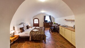 Un dormitorio acogedor con cama, mesita de noche y ventana. Se ve un área de cocina con pileta, placares y una mesa de comedor a través de un arco.