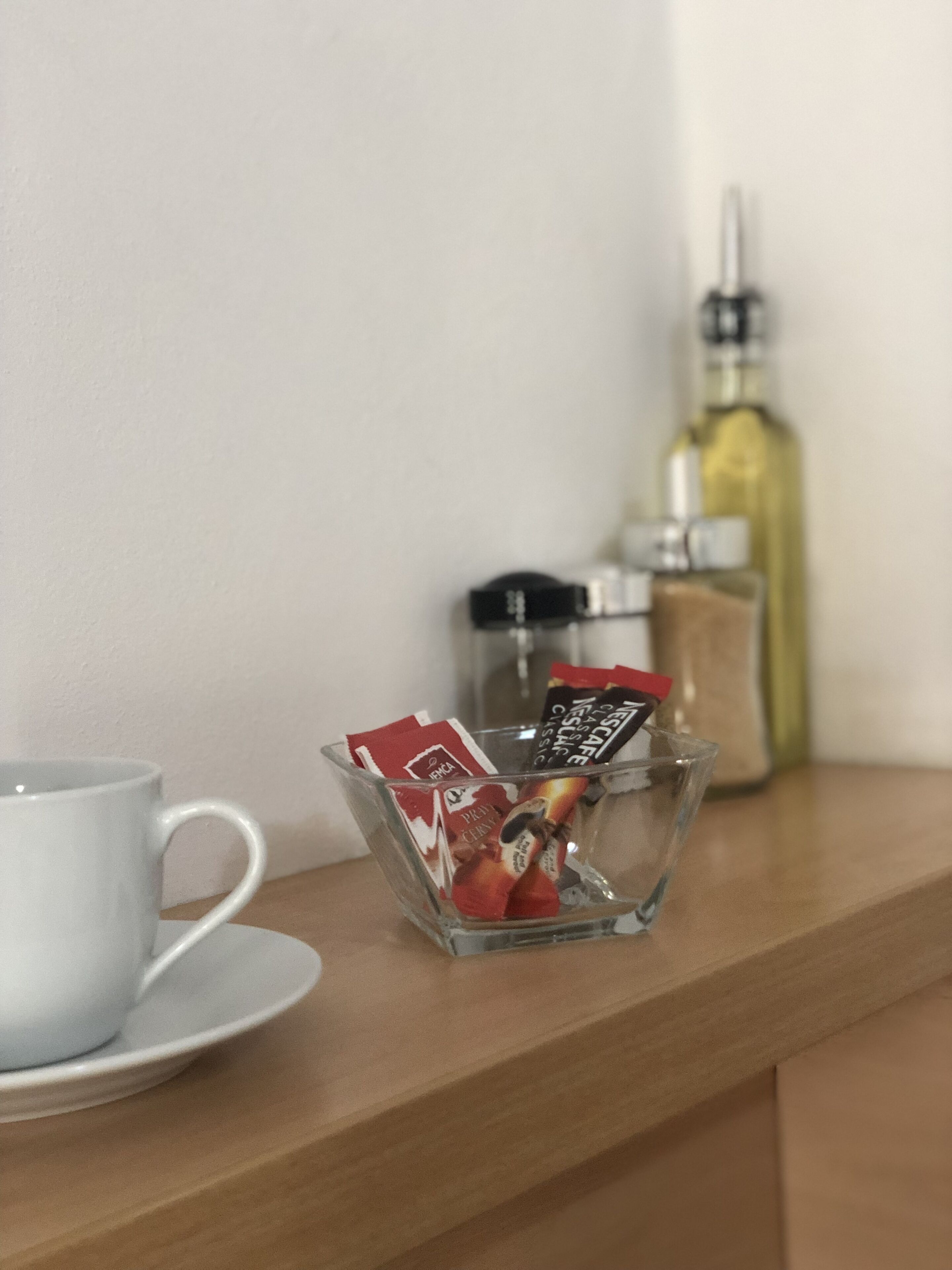 Una tassa de cafè sobre un safata, un bol de vidre amb càpsules de cafè i una ampolla d'oli d'oliva damunt una superfície de fusta.