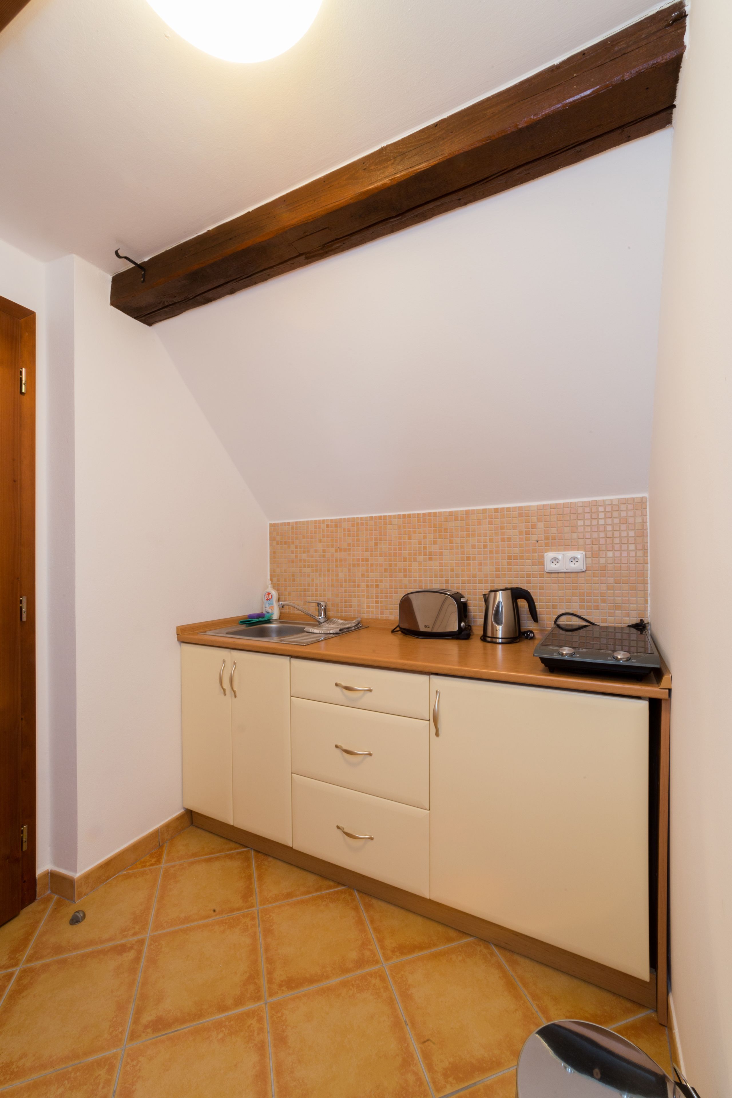 A small kitchen with a tiled backsplash, a wooden beam ceiling, and a sink with a kettle and toaster on the counter.