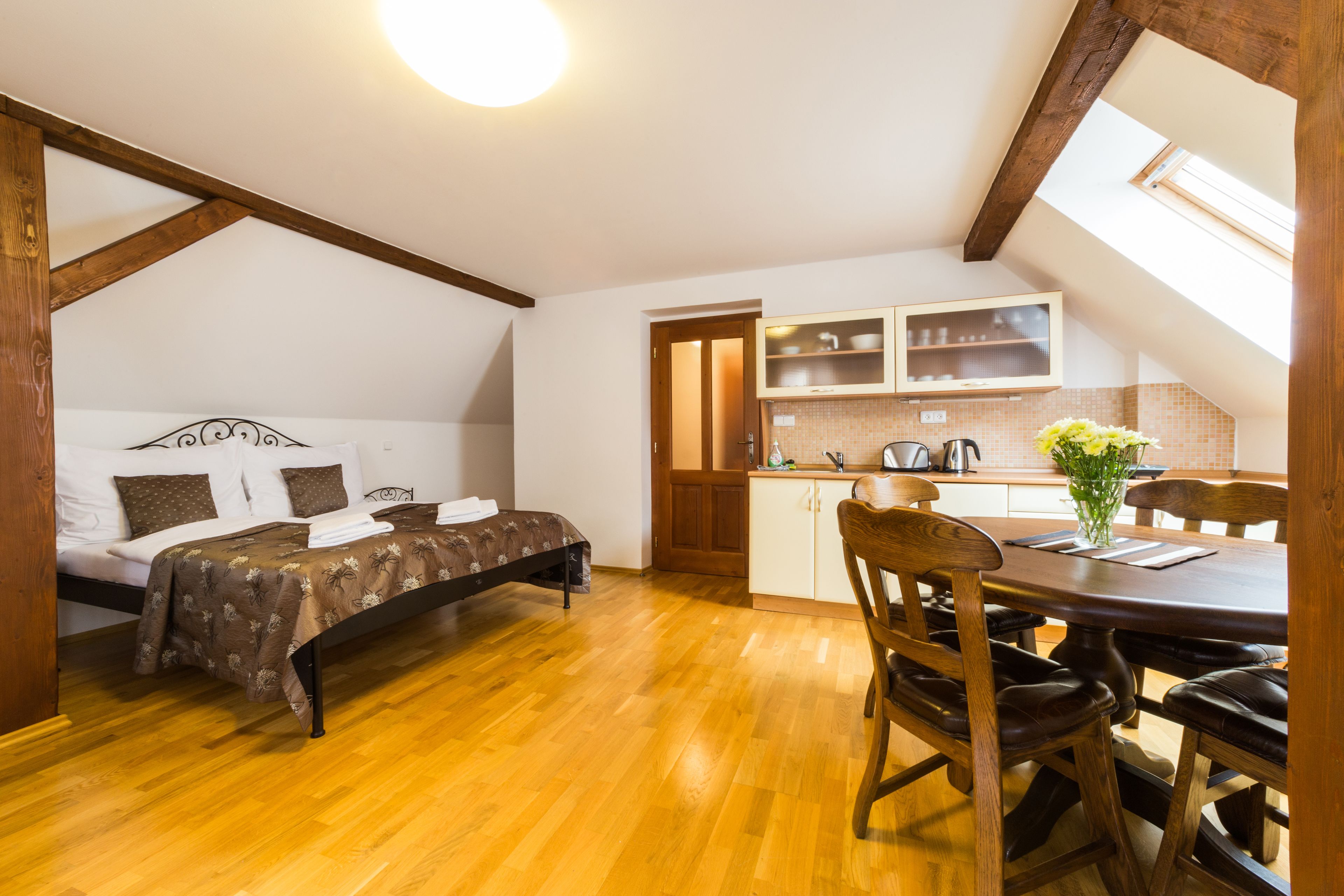 A bedroom with a bed, a wooden floor, a skylight, a kitchenette, and a dining area with a table and chairs.