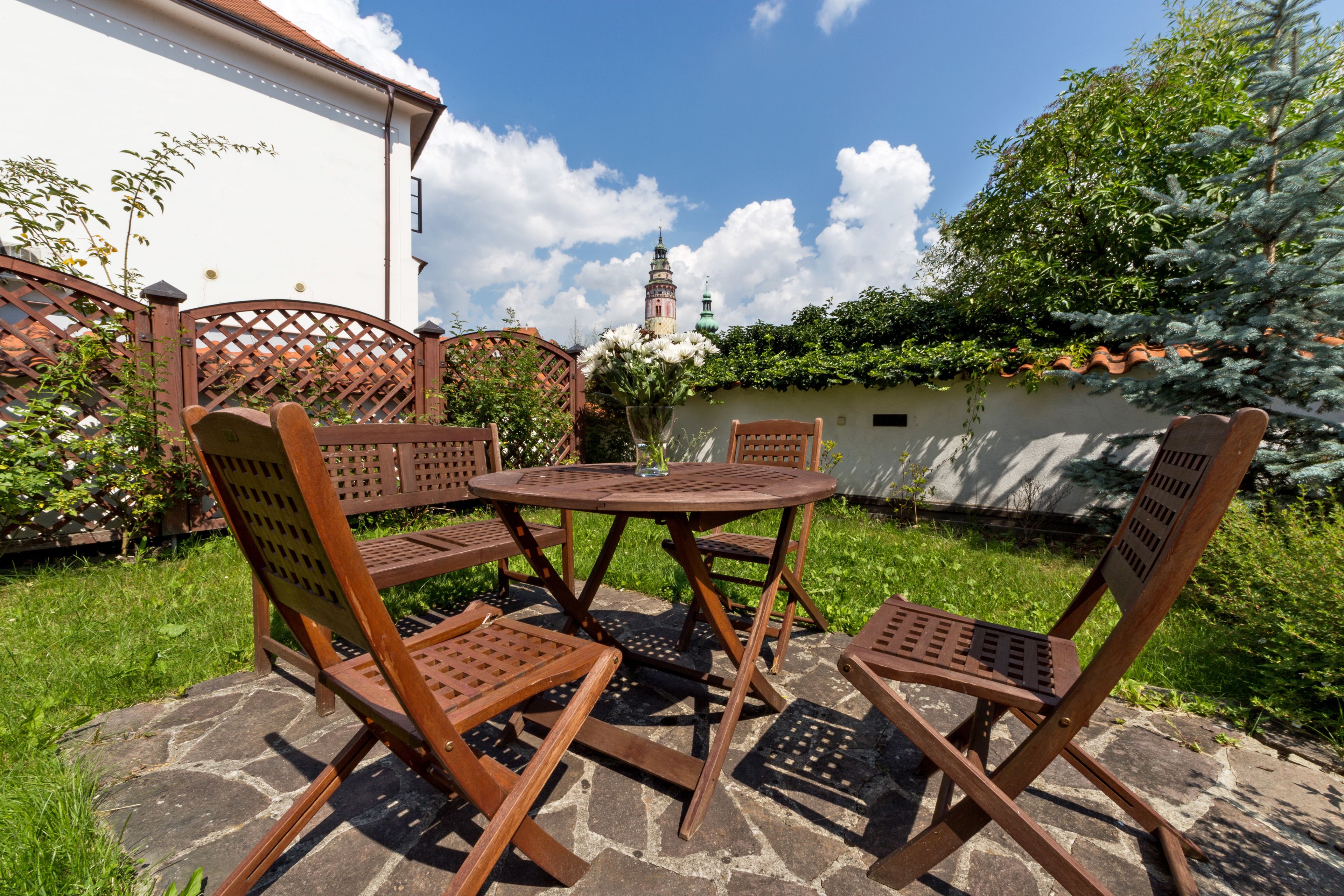 An outdoor patio with a round table and four wooden chairs, surrounded by a stone floor and lush greenery.