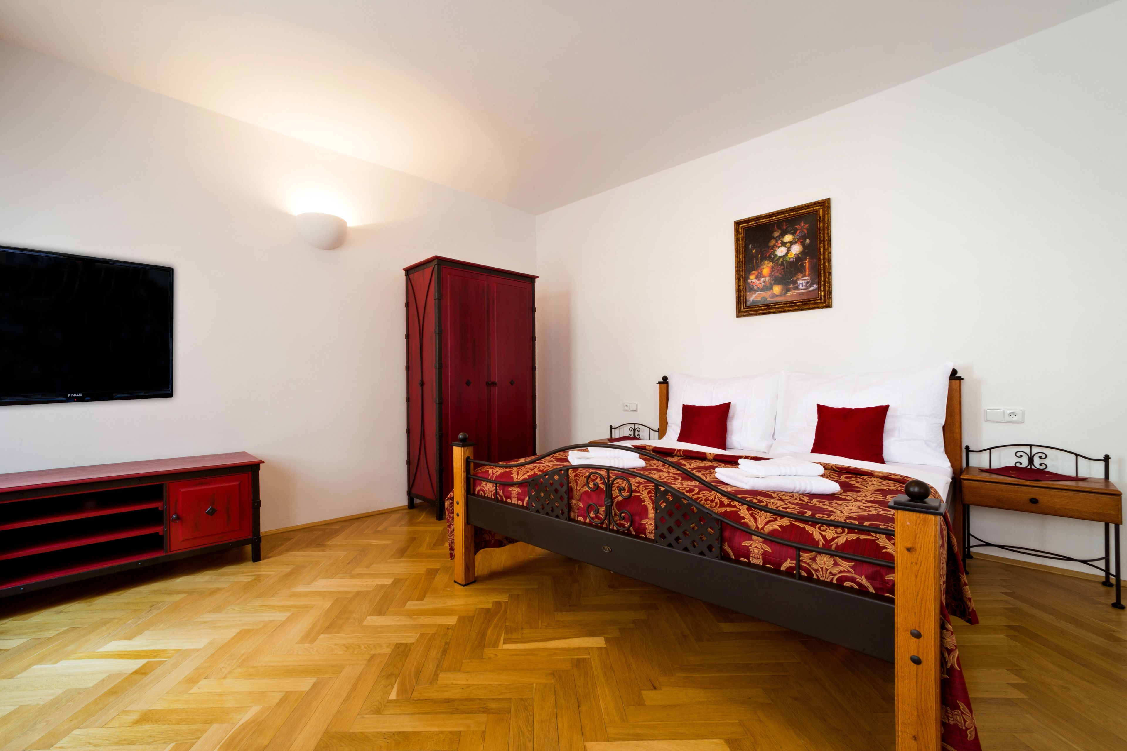 Un dormitorio con suelo de madera, cama con ropa de cama roja y blanca, un armario rojo, un televisor en la pared y un cuadro sobre la cama.