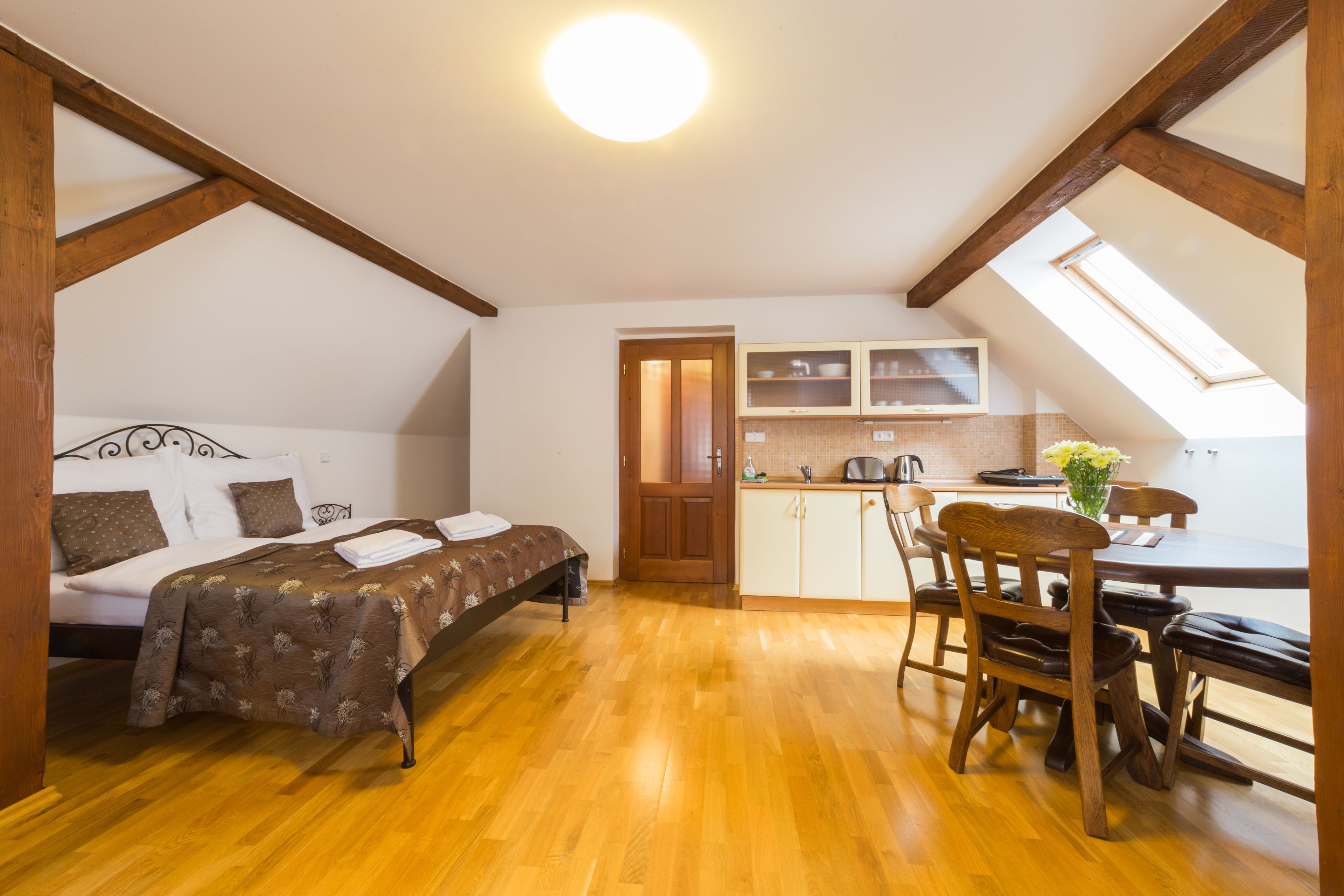 A room with a bed, wooden floor, a dining table with chairs, a kitchen area with cabinets, and a skylight.