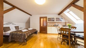 A room with a bed, wooden floor, a dining table with chairs, a kitchen area with cabinets, and a skylight.
