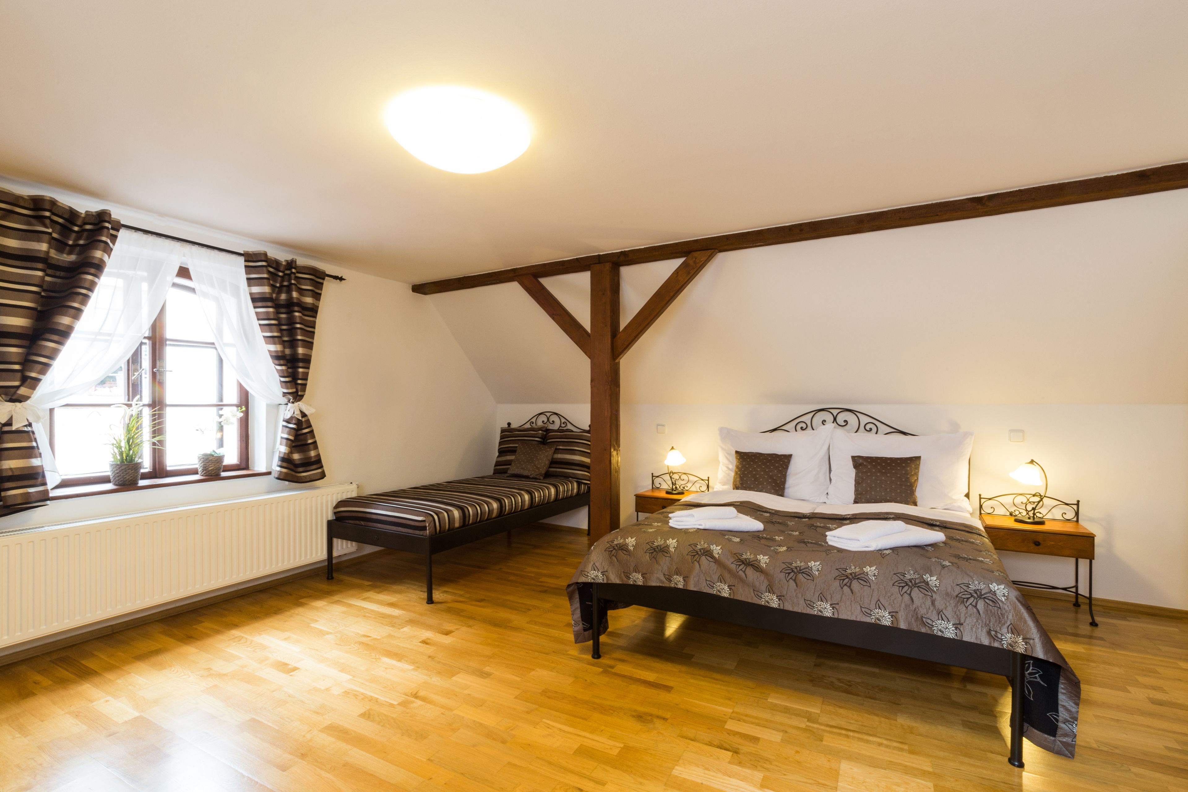 Une chambre avec un grand lit, un lit plus petit, une fenêtre avec des rideaux, un radiateur et une poutre en bois au plafond.