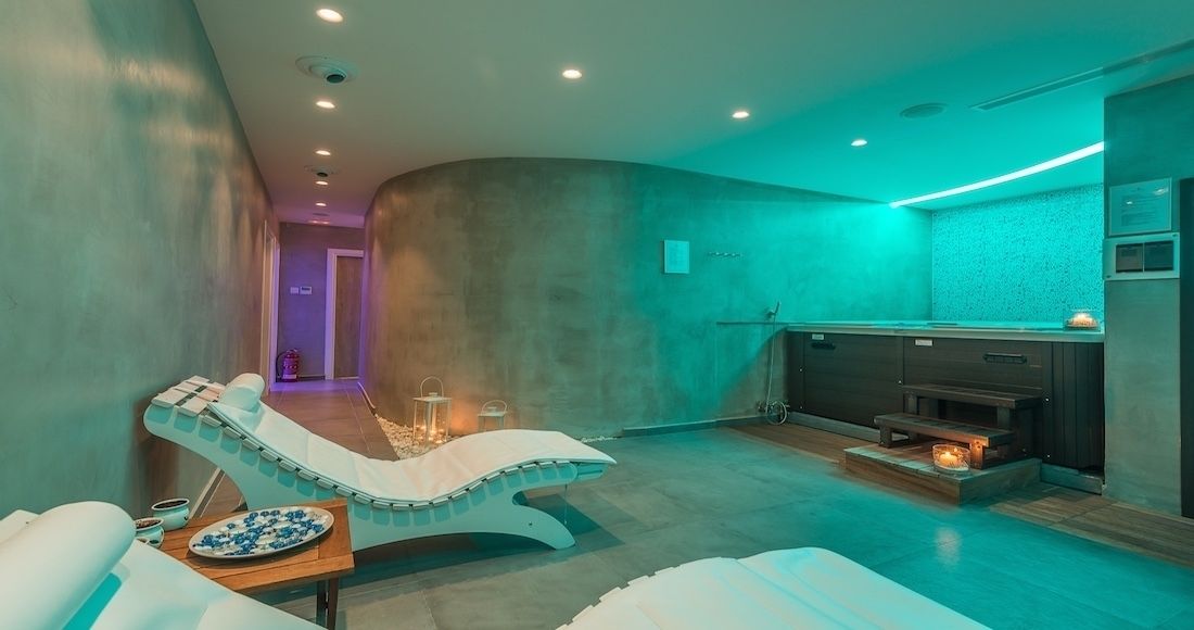 A modern spa room with a curved wall, a massage chair, a small table with a bowl of fruit, and a fireplace.