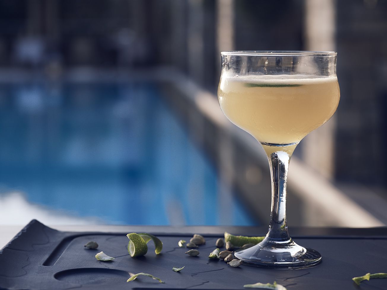 A cocktail glass with a clear liquid on a dark surface near a pool.