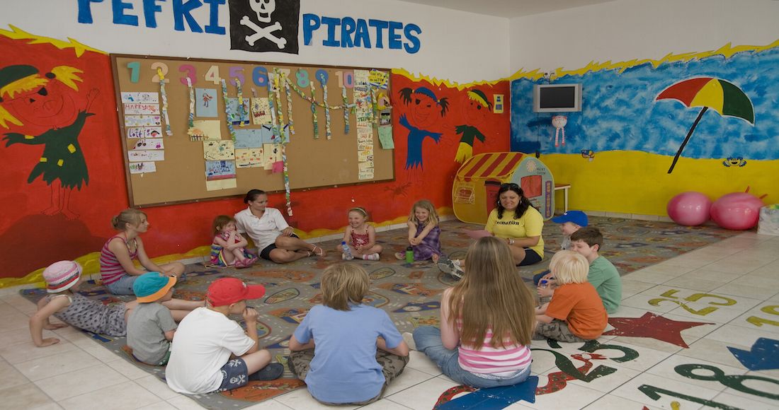 Un aula con niños sentados en el suelo, un tablón de anuncios con trabajos infantiles y una pared decorada con temática pirata.