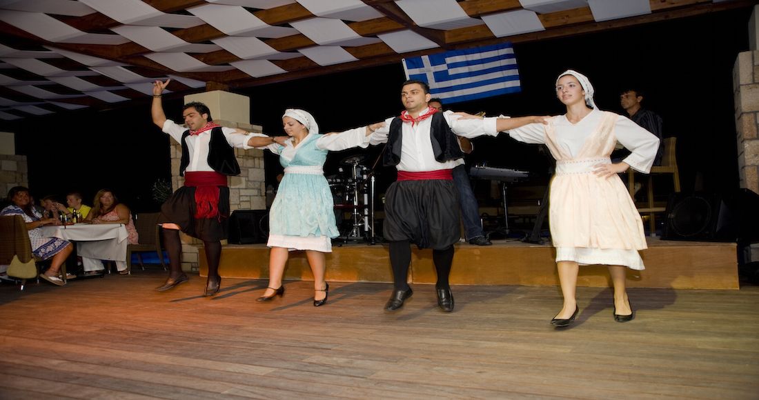 Een groep die een traditionele dans uitvoert op een podium met een houten vloer en een Griekse vlag op de achtergrond.