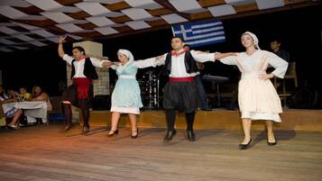 Un grupo interpreta una danza tradicional en un escenario con suelo de madera y una bandera griega al fondo.