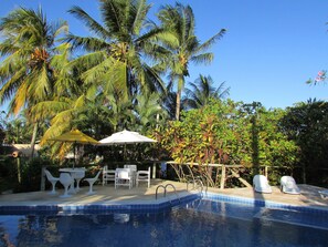 Una piscina con bordo piastrellato, circondata da palme e un patio con sedie e ombrelloni bianchi.