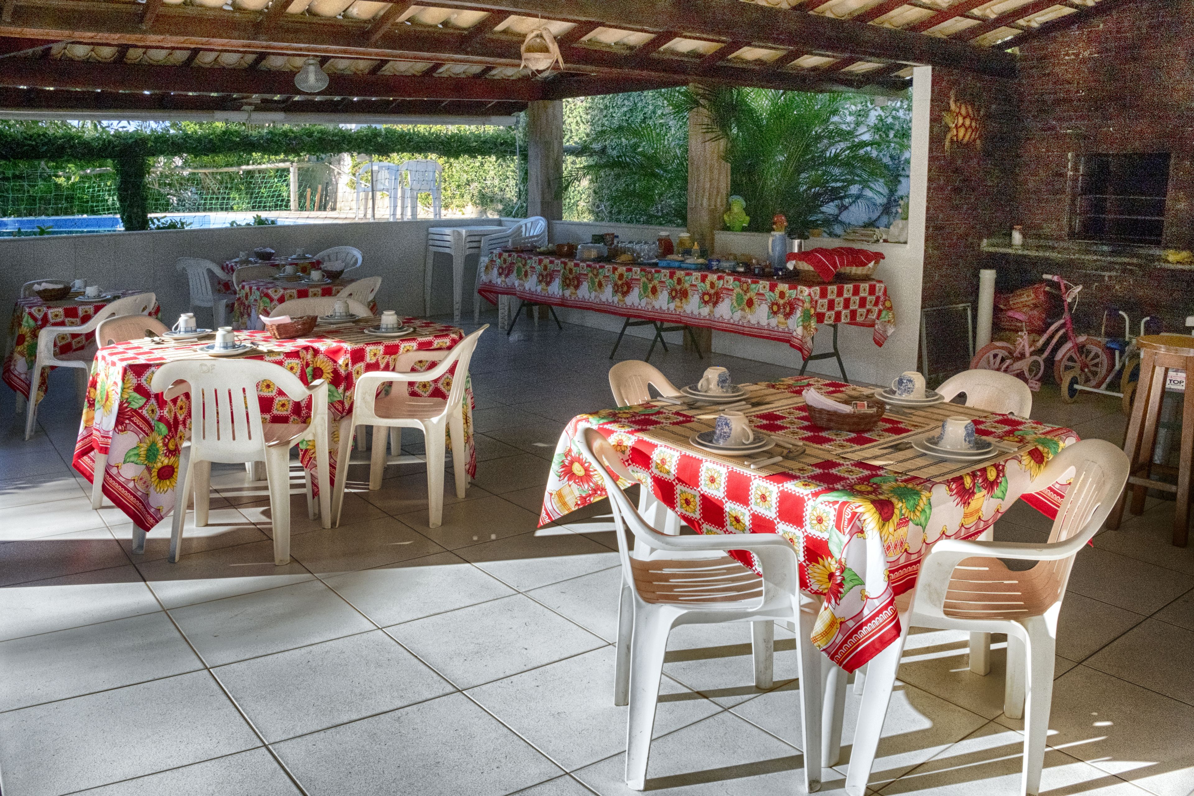 An outdoor dining area with multiple tables covered with red and white checkered tablecloths, surrounded by white plastic chairs.