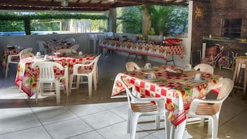 An outdoor dining area with multiple tables covered with red and white checkered tablecloths, surrounded by white plastic chairs.