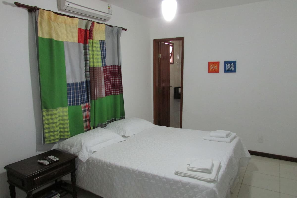 Un dormitorio con una cama, una mesita de noche con libros, una cortina colorida y una puerta.