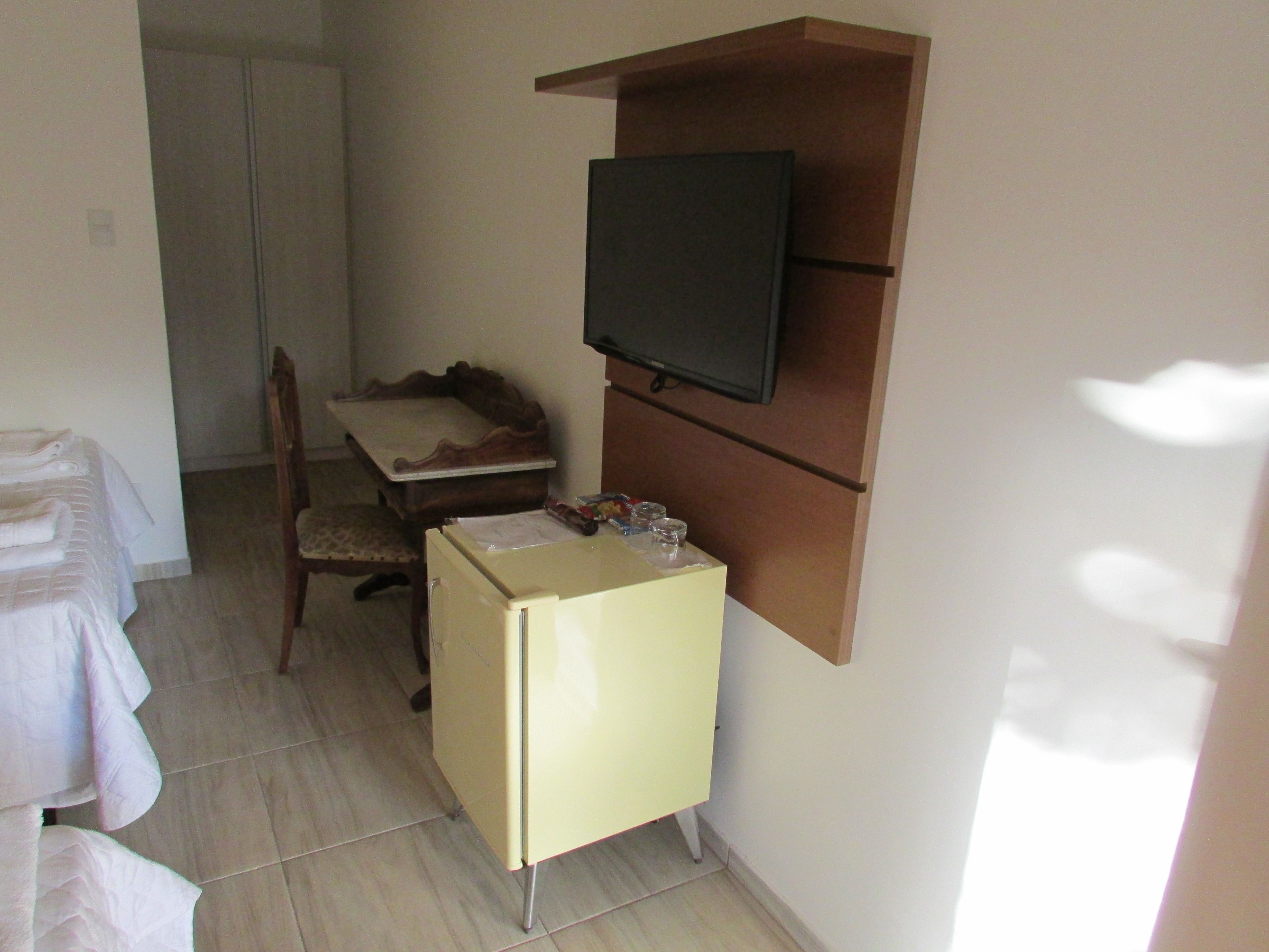 Ein kleines Zimmer mit Bett, Schreibtisch, Stuhl, einem an einer Holzablage montierten Fernseher und einem kleinen Kühlschrank.