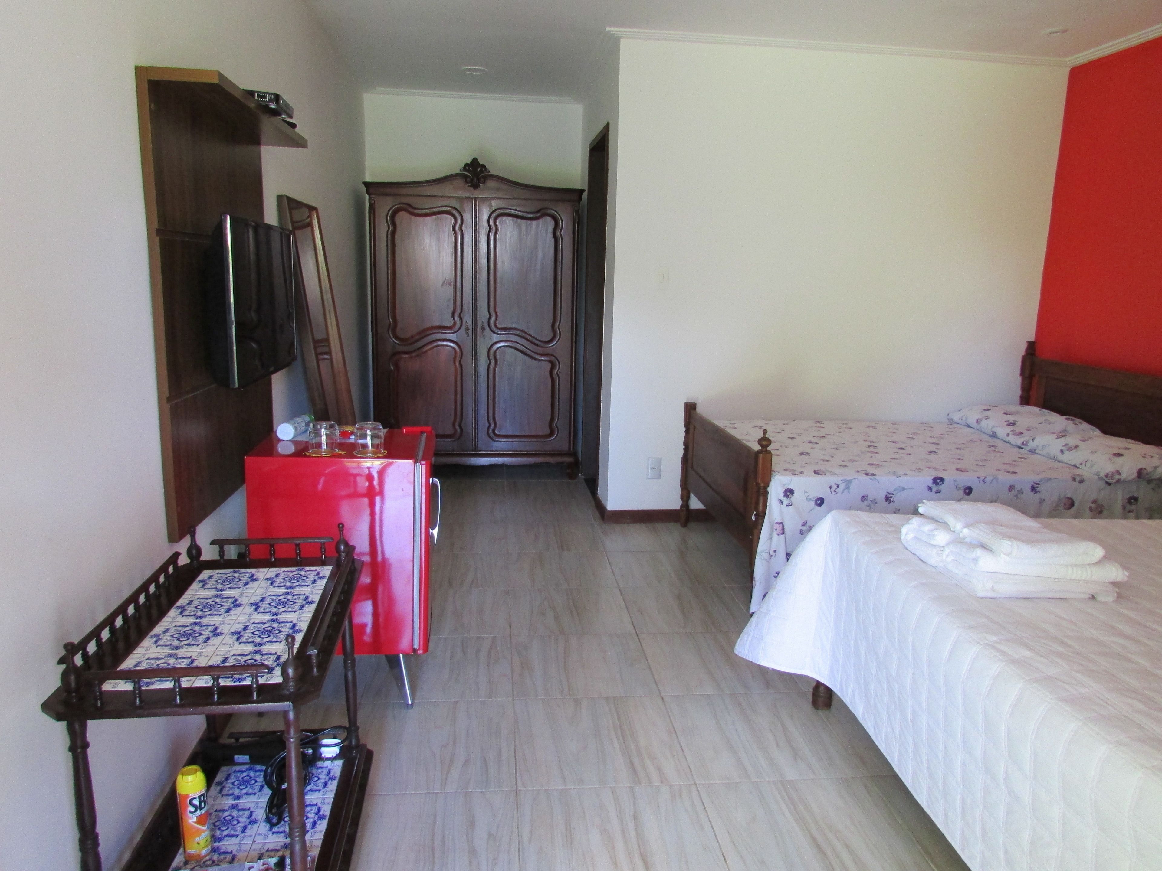 Quarto de hotel com uma cama, uma escrivaninha com cadeira, um guarda-roupa e uma televisão na parede.