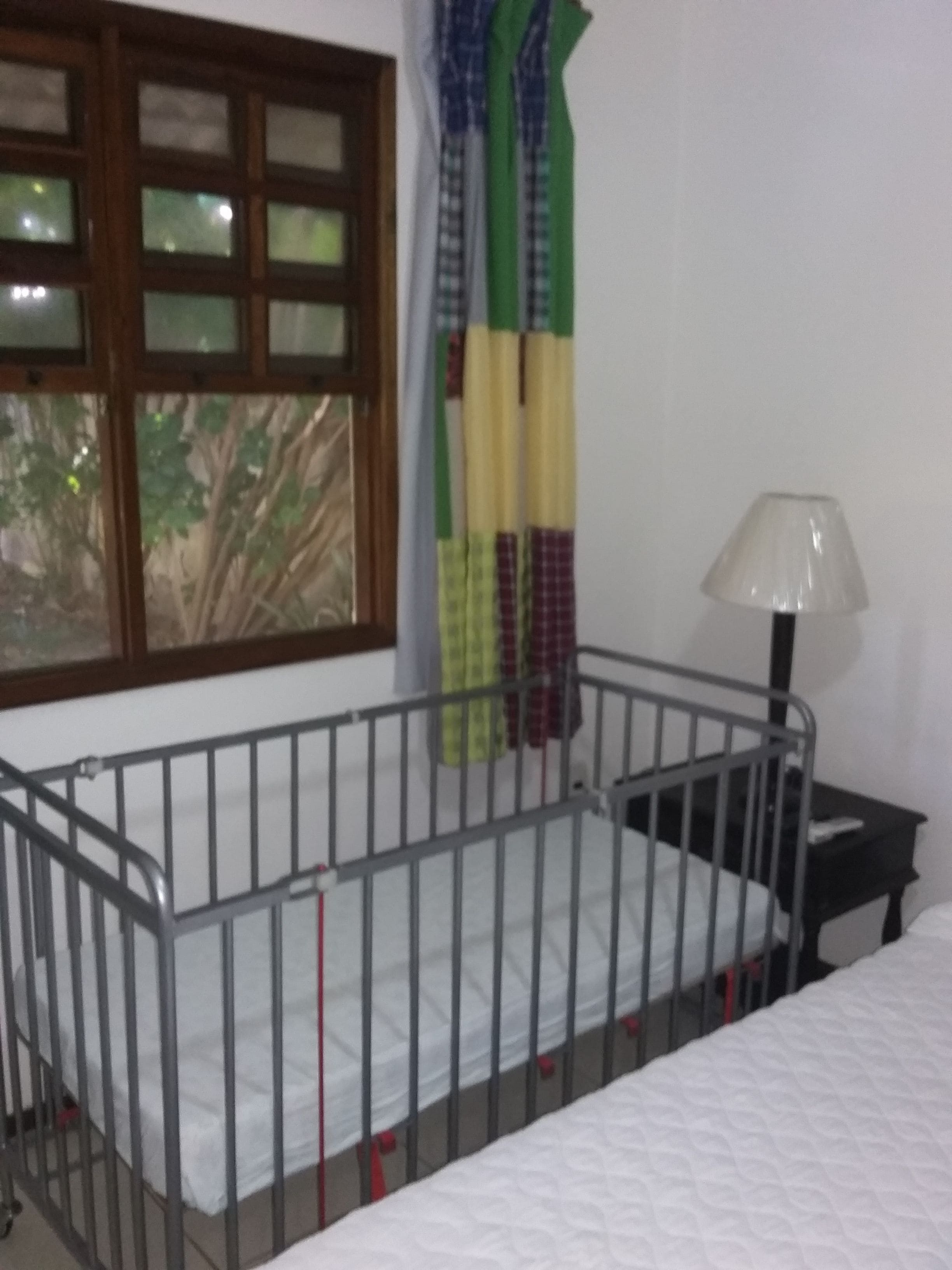A room with a crib, a window with curtains, a bedside table, and a lamp.