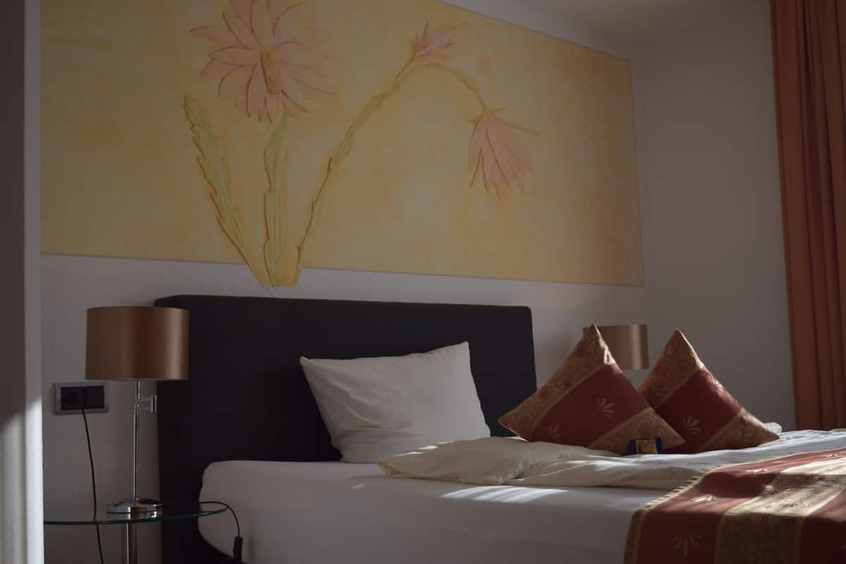 Ein Hotelzimmer mit einem Bett, zwei Nachttischlampen, einem großen Blumenbild an der Wand und einem Fenster mit Vorhängen.