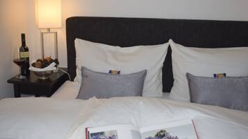 Uma cama bem arrumada com roupa branca, uma mesinha de cabeceira com uma garrafa de vinho e uma fruteira, e um abajur.