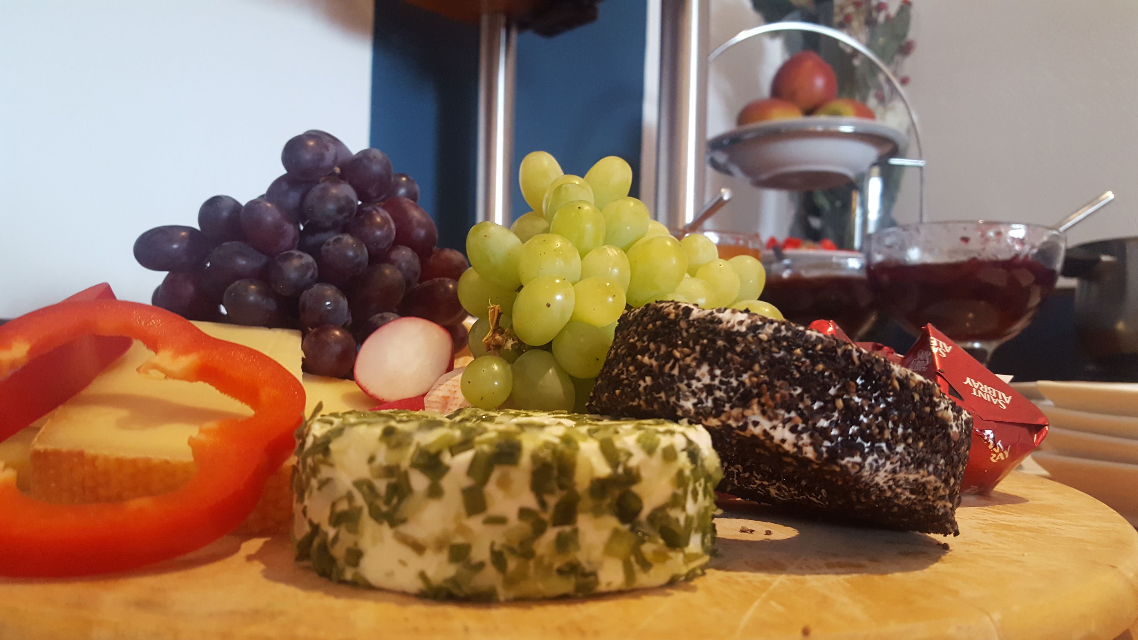 A wooden board with various cheeses, grapes, and a red bell pepper.