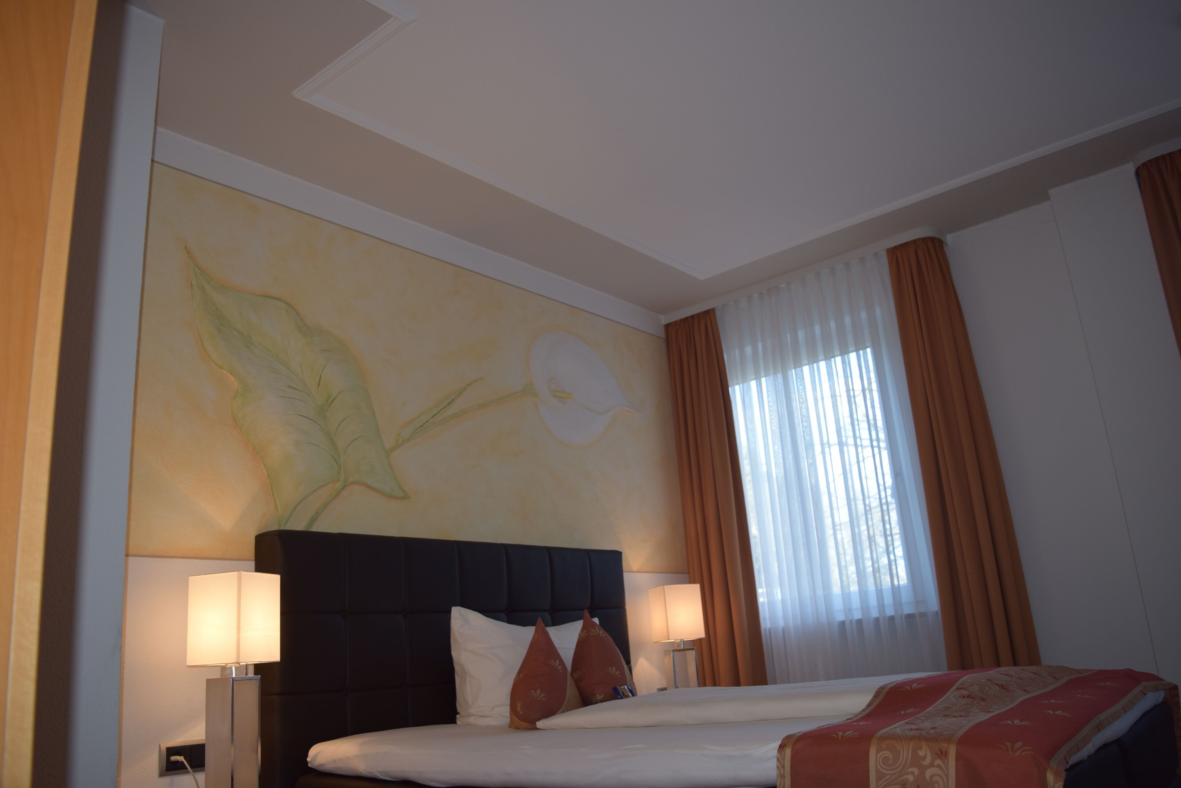 Una habitación de hotel con una cama grande, dos lámparas de noche, un mural en la pared y una ventana con cortinas.