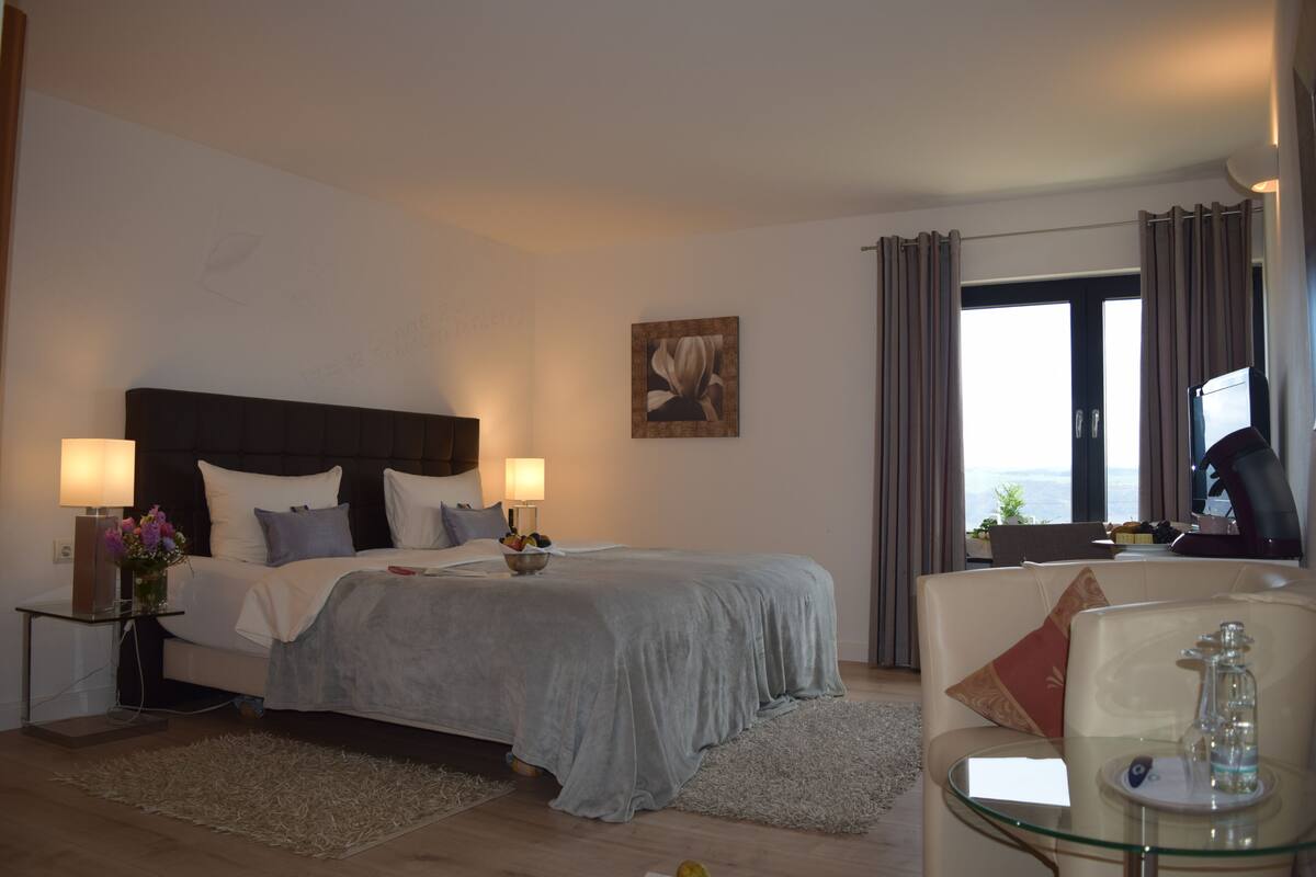 Ein Hotelzimmer mit Bett, Nachttischlampen, einem Nachttisch, einem Glastisch mit Blumen und einer Couch, mit Blick ins Freie.