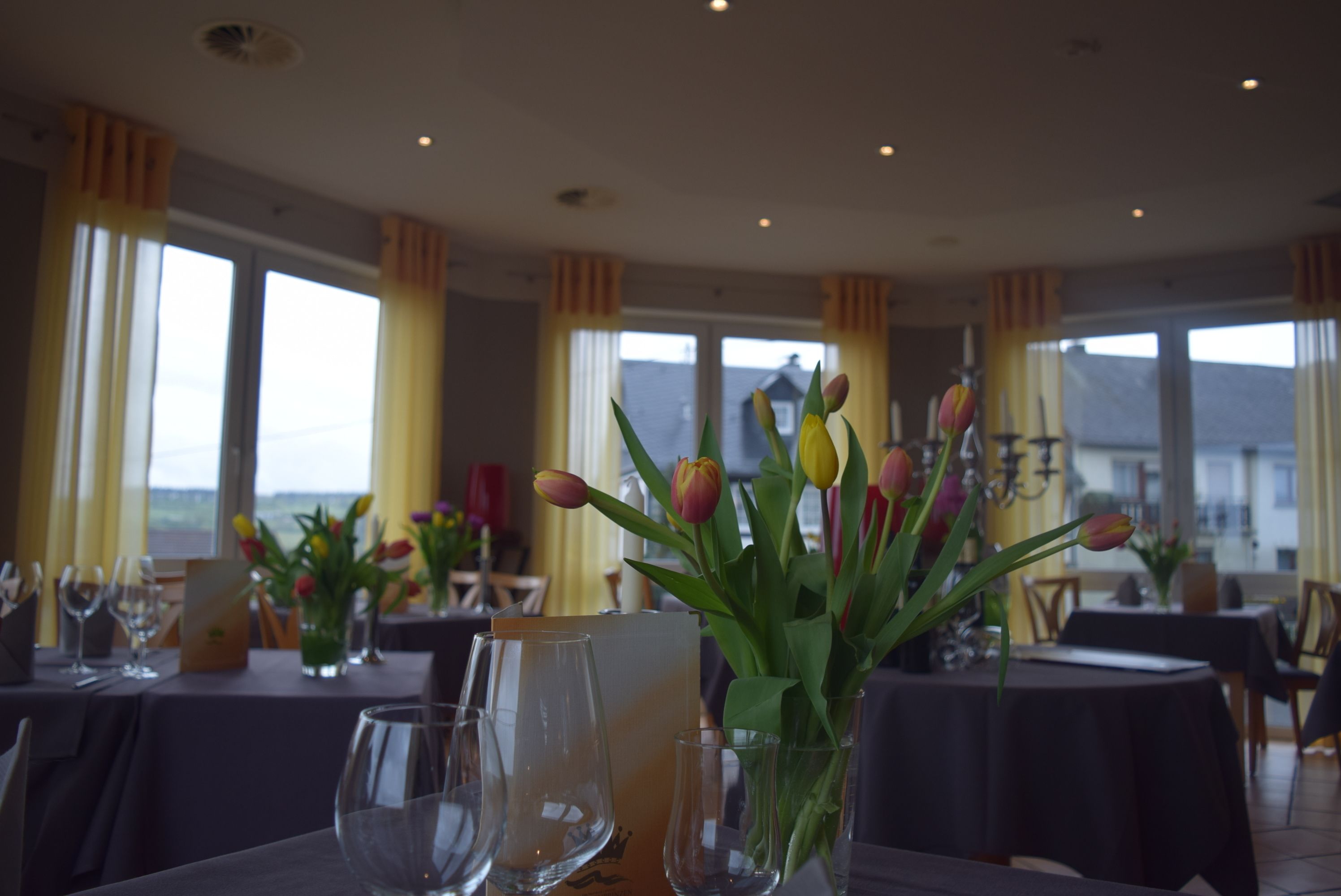 A restaurant with tables set for dining, featuring tulips in vases and a view of the outdoors.